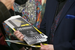Uśmiechnięty Robert Brzęcki patrzy na trzymane w prawej dłoni książki zawierające zdjęcia wystawionych w ramach "The Present" prac.