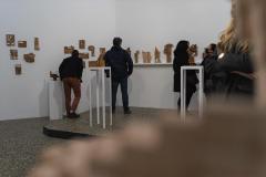 Trzy odwrócone plecami osoby oglądają drewniane rzeźby. Prace wyeksponowane są na półce i białej  ścianie