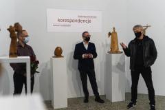 Pośród rzeźb stoją od lewej: Stanisław Kośmiński, Robert Brzęcki. Antoni Grabowski. Mężczyźni rozmawiają i mają na twarzach maseczki. W tle wyświetlony jest tutuł wystawy: Korespondencje.