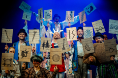 Grupa aktorów z tabliczkami z rysunkami umieszczonymi na patykach.
