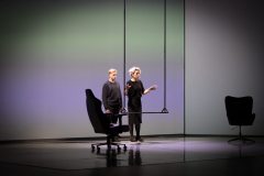 W środku kadru Katarzyna Herman i Konrad Szymański na scenie. Przed niimi huśtawka. Po bokach dwa ciemne fotele obrotowe.