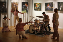 Cztery tancerki w układzie z różową włóczką. Między nimi perkusista. W głębi kadru prace fotograficzne wyeksponowane na białej ścianie i jasnej kotarze.
