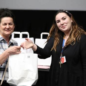 Nikola Dębowska z CKiS w Konine wręcza białą torebkę z numerem 10 kobiecie w koszuli w kartkę. Za nimi papierowe torebki na brzegu sceny.