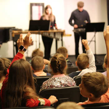Zdjęcie wykonane zza rzędów krzeseł, na których siedzą dzieci. Niektóre podnoszą ręce do góry. W tle muzycy na scenie.