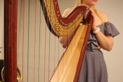 Kobieta w długiej sukni z odkrytymi ramionami opiera dłonie o harfę. Uśmiecha się.