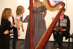 W centrum kadru harfa stojąca na środku małej sceny, a za nią kobieta w długiej sukni z odkrytymi ramionami. Obok stoi dziewczynka. Przed sceną kobieta w czerni z kręką opartą na biodrze i mikrofonem.