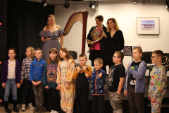 Dzieci stoją w rzędzie przed sceną pozując do zdjęcia. Na scenie trzy kobiety. Jedna z nich trzyma waltornię. Obok stoi harfa.