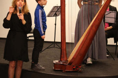 Przed sceną kobieta w czerni mówi do mikrofonu. Na scenie chłopiec przy harfie. Za instrumentem kobieta w długiej sukni.