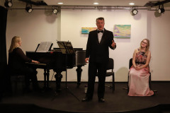 Trio na scenie. Od lewej: siedząca przy fortepianie kobieta w czerni, śpiewający mężczyzna w garniturze, siedząca na krześle kobieta w kolorowej sukience przechylona w bok.