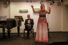 Kobieta w długiej kolorowej sukience śpiewa. Podnosi ręce. Za nią na krześle siedzi mężczyzna. Po lewej pulpit z nutami i fragment fortepianu.