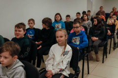 Dzieci siedzące przy ścianie na krzesłach ustawionych w rzędy. W drugim rzędzie dziewczynka w białej bluzie z napisami patrzy w obiektyw i się uśmiecha.