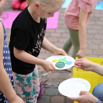 W środku kadru dziewczynka z papierowym talerzykiem, na którym są barwne kleksy farb.