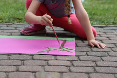 Dziewczynka maluje drzewo na różowym brystolu. Na pierwszym planie rozmazany pęk pędzli.