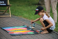 Kucająca na chodniku dziewczynka w białej koszulce, ciemnych leginsach i czapce z daszkiem podczas malowania pracy. W tle para nóg.