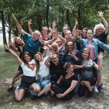 Zdjęcie grupowe uczestników warsztatów, machających do fotografa. Polanka pośród drzew.