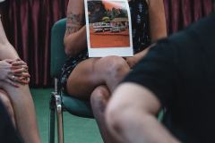 Siedząca na krześle kobieta z tatuażem prezentuje reprodukcję, na której widać dwa samochody. Po lewo noga innej uczestniczki, po prawo łokieć i rękaw czarnej koszulki.