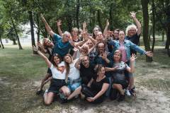 Zdjęcie grupowe uczestników warsztatów, machających do fotografa. Polanka pośród drzew.