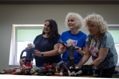 Trzy uśmiechnięte kobiety stoją przy stole z marionetkami. W tle głowa kucającej kobiety i dwa okna.