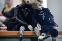 Zbliżenie na dwie marionetki siedzące na brzegu stolika. Czarne kostiumy, zwierzęce głowy.