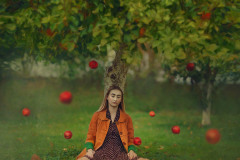 Pod drzewem w rozkroku siedzi modelka w pomarańczowej kurtce i sukience w groszki. Wokół niej latające jabłka.