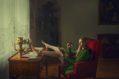 Na czerwonym fotelu przy oknie siedzi modelka w zielonym swetrze i sukience w groszki. Gołe nogi trzyma na blacie. Przez lupę studiuje książkę.