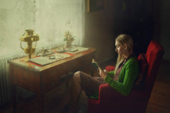 Na czerwonym fotelu siedzi modelka ubrana w zielony sweter i sukienkę w groszki. Czyta książkę. Przed nią biurko ustawione przy oknie. Na blacie rozłożone dokumenty, lupa i lampa naftowa.