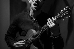 Mężczyzna gra na gitarze i śpiewa. Ma szeroko otwarte usta.  Zdjęcie czarno-białe.