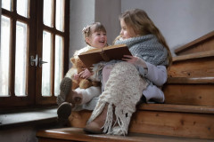 Dwie dziewczynki siedzą na stopniach drewnianych schodów. Trzymają otwartą książkę. Po lewej okno.
