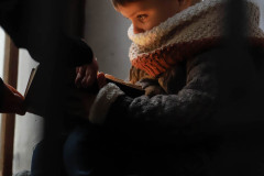 Chłopiec siedzący na stopniu schodów z książką. Sfotografowany z lewego profilu. Książkę łapią czyjeś dłonie.