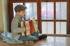 Chłopiec z akordeonem siedzi na drewnianej podłodze przy oknie. Ubrany w szary sweter i niebieskie dżinsy. Na głowie kaszkiet.