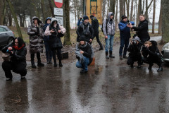Grupa fotografów na mokrym od deszczu asfalcie. Część przyklęka i robi zdjęcia. W tle dwa zaprakowane samochody, znaki drogowe i pnie drzew.
