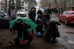 Deszczowa pogoda. Grupa fografów w trakcie robienia zdjęć. Część osób kuca. Jedna ukrywa się pod zielonym parasolem.