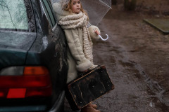 Dziewczynka opiera się o samochód. W prawej ręce trzyma starą walizkę podróżną, w lewej przezroczysty parasol chroniący ją przed deszczem. Sfotografowana z prawego profilu.