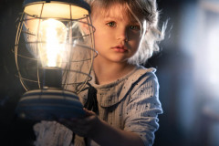 Dziecko trzymające za uchwyt lampkę z zapaloną żarówką patrzy w obiektyw. Ubrane w sukienkę w kropki. Włosy blond.