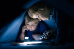 Dwoje dzieci czyta książkę pod kocem. W dłoniach trzymają zapalone latarki.