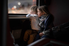 Dwoje przytulonych dzieci patrzy przez okno pociągu. Jedno z nich opiera ramiona na stoliku przy oknie. Obok na siedzeniu leży stara walizka.