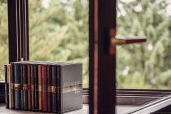 Książki autorstwa Ryszarda Kapuścińskiego ustawione w równym rzędzie. Ułożone na parapecie przy szeroko otwartym oknie. W tle zieleń drzew.