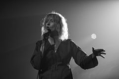 Mela Koteluk śpiewa. W prawej ręce trzyma mikrofon, lewą unosi na wysokość pasa. Zdjęcie czarno-białe.