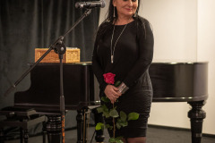 Iza Kostiukow w czarnej sukience z długą czerwoną różą stoi przy mikrofonie. Wzrok skierowany w dół. W tle fortepian i ciemne kotary.