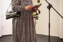 Kobieta w długiej sukni w kwiatowy deseń stoi przy mikrofonie. W dłoniach trzyma różę i katalog wystawy.