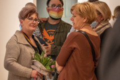 Chwila rozmowy podzcas wernisażu. Po lewej kobieta w beżowym swetrze trzymająca bukiet tulipanów, po prawej kobieta w brązowym sweterku z czarnym paskiem torebki. Pośrodku mężczyzna z dzieckiem na ręku. Na twarzy tęczowa maska.