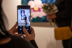 Na pierwszym planie plecy i ręce osoby trzymającej telefon. Na wyświetlaczu kobieta pozuje do zdjęcia przy jednej z prac.