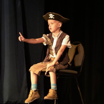 Chłopiec w stroju pirata siedzi na krześle. Podnosi w górę prawą rękę z uniesionym palcem.