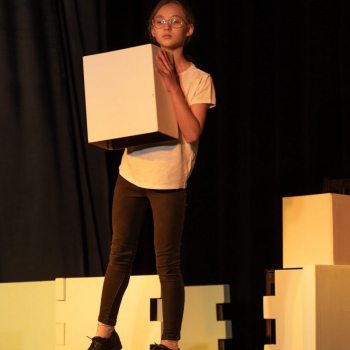Dziewczynka w okularach, białej koszulce i ciemnych spodniach trzyma przed sobą białe pudełko. Za nią kartony i inne elementy układanki.