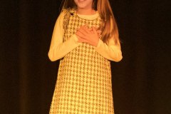 Dziewczynka uczesana w dwie kitki, ubrana w sukienkę w pepitkę przyciska dłonie do serca.