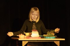 Dziewczynka z blond włosami ściętymi do ramion rozkłada ręce nad stolikiem, na którym leżą naczynia kuchenne, imitacja toryu, packa i motyl.