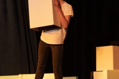 Dziewczynka w okularach, białej koszulce i ciemnych spodniach trzyma przed sobą białe pudełko. Za nią kartony i inne elementy układanki.