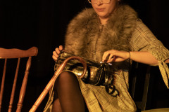 Dziewczyna w okularach, kraciastej sukience i futrzanym szalu siedzi na krześle. Na kolanach trzyma torebkę i opiera laskę.
