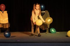 Dziewczynka z długimi włosami spiętymi w dwie kitki trzyma dwa balony. Obok leżą inne kolorowe balony. W tle dwa krzesła obleczone w ubrania.