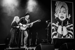 Po lewej Sylwia Dziardziel za plecami gitarzysty. Po prawej portret Kory Jackowskiej. Zdjęcie czarno-białe.
